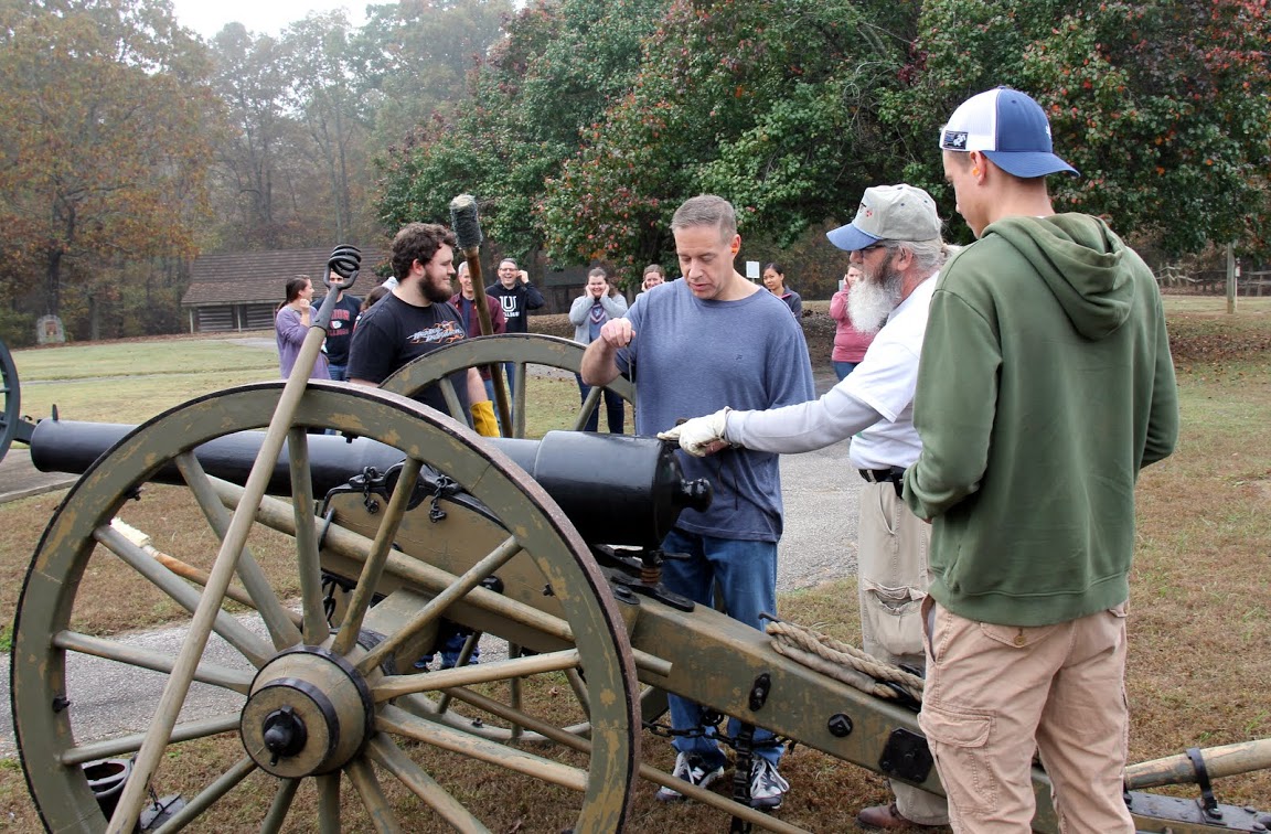 Dr. Henry Allen (foreground, second from left) inserts a priming wire down the cannon vent to prepare a powder packet for ignition during the Phi Alpha Theta-sponsored community service project at Britton Lane Battlefield on November 3, 2015. Others in the foreground are Nicholas Hancock (left), James Price (second from right), and Stephen Lewoczko (right, back to camera).