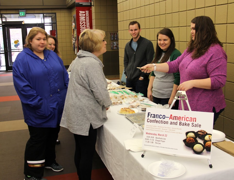 Linda Taylor (wearing gray jersey coat) buys some cinnamon rolls from Jessica Barton (right) at the Phi Alpha Theta bake sale on March 22, 2017, as salespersons Seth Reid (third from right) and Jordan Sellers (second from right) look on.