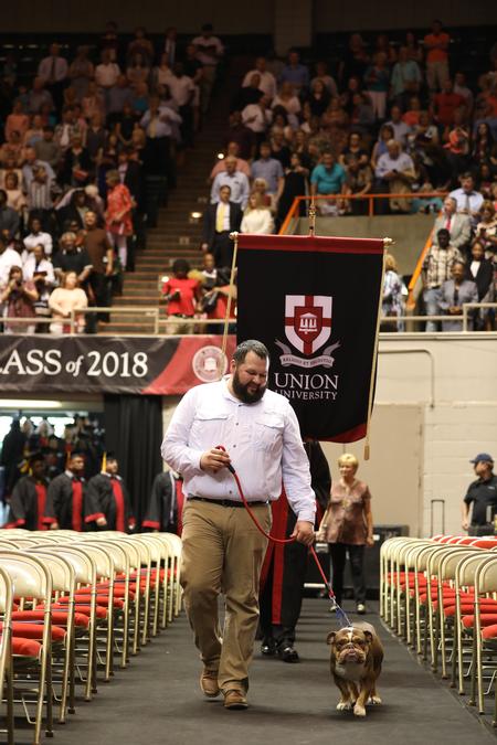 Rusty Tuders, assistant resident director, walks Buster the Bulldog down the aisle as the graduates process into the 2018 spring commencement services. - Photo by Kristi McMurry Woody