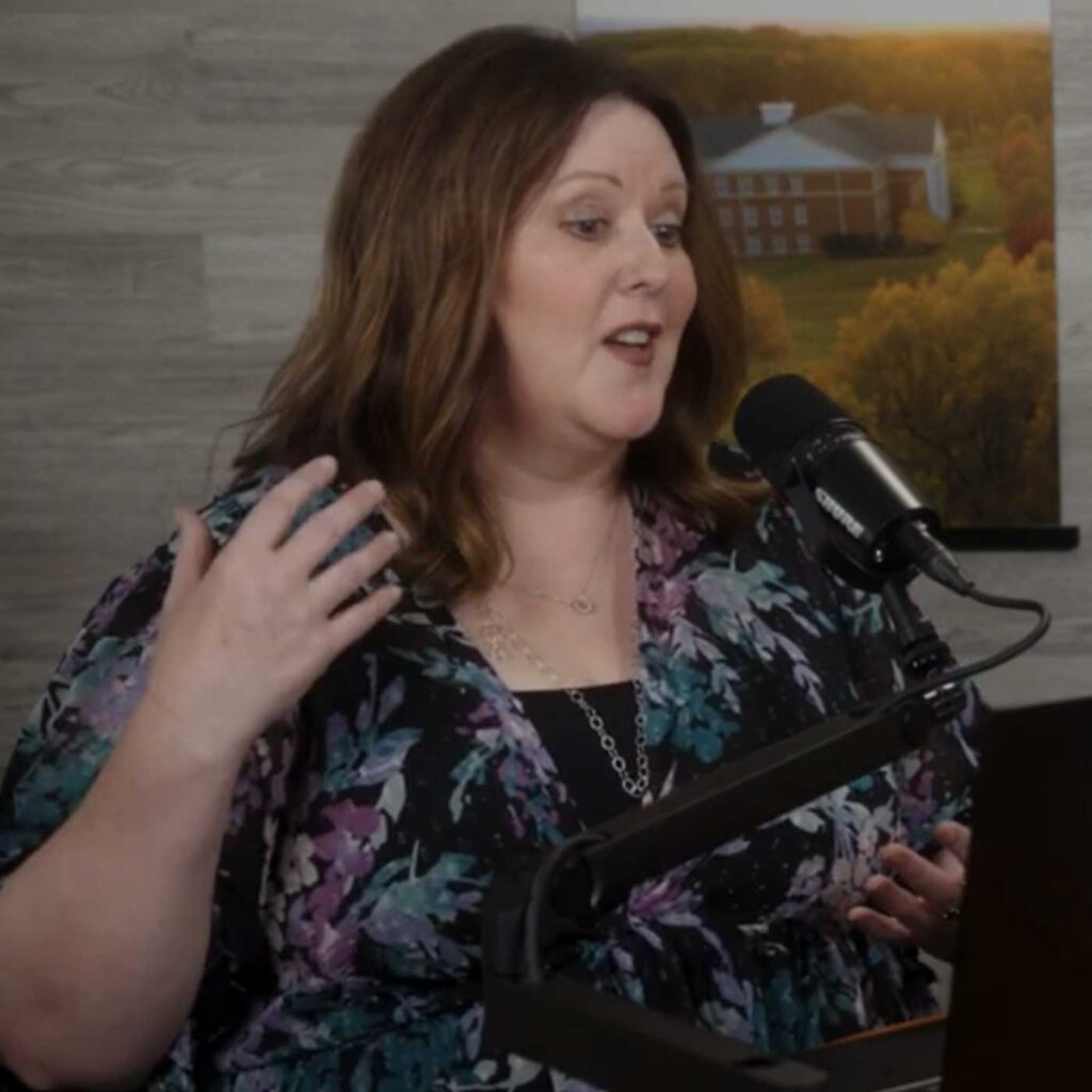 A woman with shoulder-length brown hair speaks into a microphone while gesturing with one hand. She is wearing a floral-patterned top and is seated indoors, with a framed landscape photo on the wall behind her.