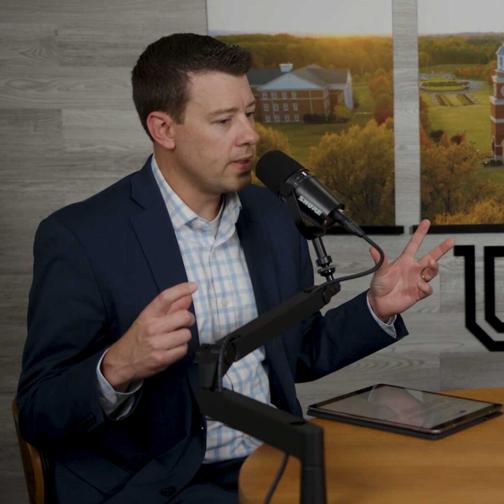 Man in a suit speaks into a microphone during a podcast or interview. He gestures with both hands, and an electronic tablet rests on the table. Framed aerial campus photos are on the wall behind him.