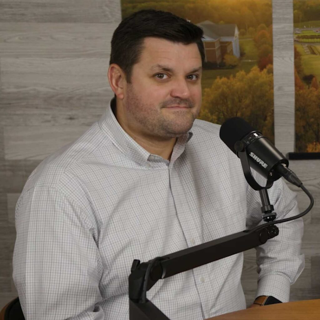 A man in a light plaid shirt sits at a desk with a microphone in front of him, positioned for a podcast or recording. The background features wooden paneling and a partially visible landscape photo.