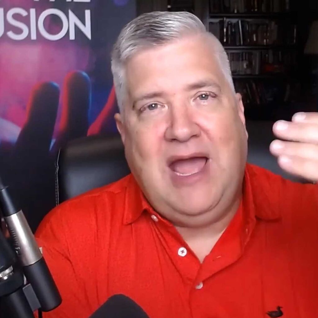 A man with short gray hair wearing a red shirt speaks into a microphone in a room with bookshelves. His right hand is raised, and there is a sign with partially visible text in the background.