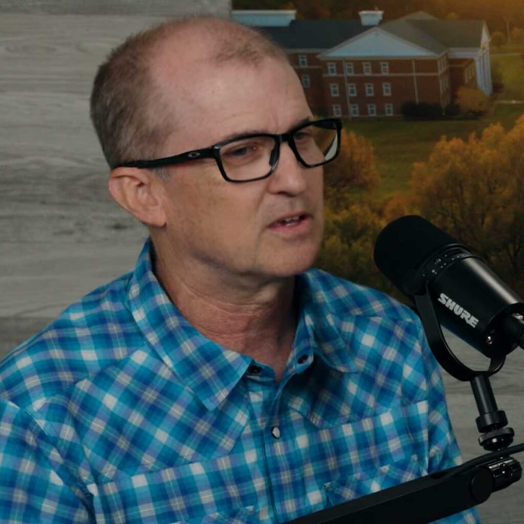 Dusty Doddridge, a middle-aged man wearing glasses and a blue plaid shirt, speaks into a studio microphone. A blurred background shows a building and trees.