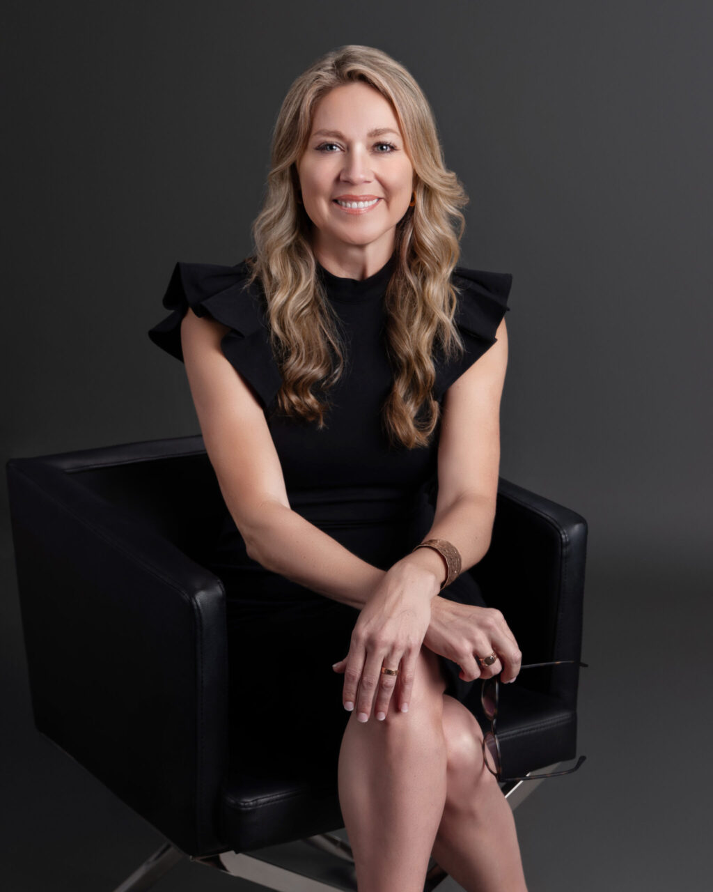 A woman with long, wavy blonde hair sits in a black chair against a dark background, wearing a black dress with ruffled sleeves. She is smiling, with her hands resting on her knee and holding eyeglasses, embodying the grace of Jill Taylor as she reflects on using gifts to serve others.