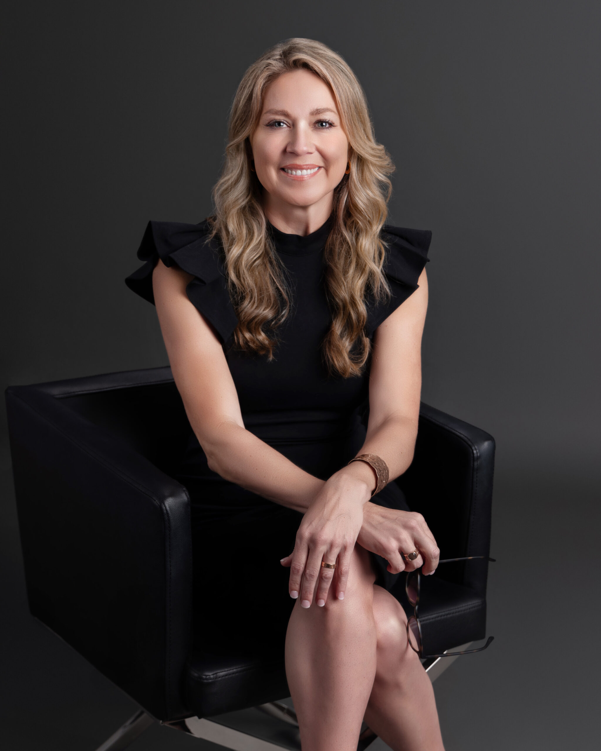 A woman with long, wavy blonde hair sits in a black chair against a dark background, wearing a black dress with ruffled sleeves. She is smiling, with her hands resting on her knee and holding eyeglasses, embodying the grace of Jill Taylor as she reflects on using gifts to serve others.