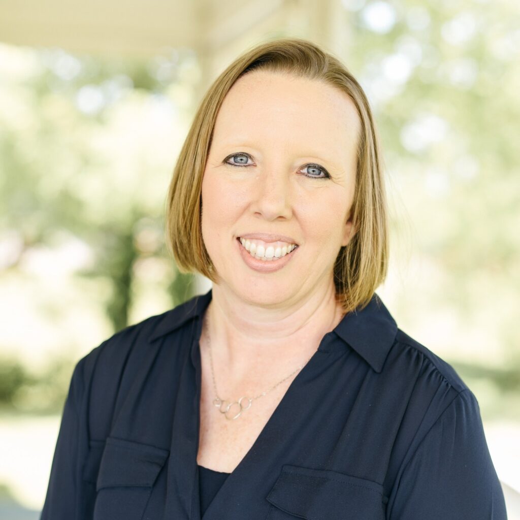 A woman with straight, light brown hair and a navy blouse smiles at the camera. She is standing outdoors in soft, natural light with a blurred green background, representing the grace and poise of Union University notable alumni.