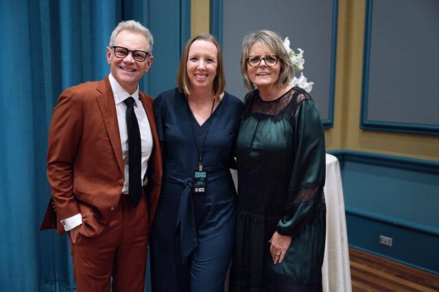 Three adults pose together indoors in semi-formal attire, exemplifying the distinguished presence of Union University Notable Alumni. The man wears a rust-colored suit and glasses, while the two women don elegant dark dresses. They stand in front of a round table adorned with a white cloth and flowers, capturing a moment of celebration among accomplished graduates.