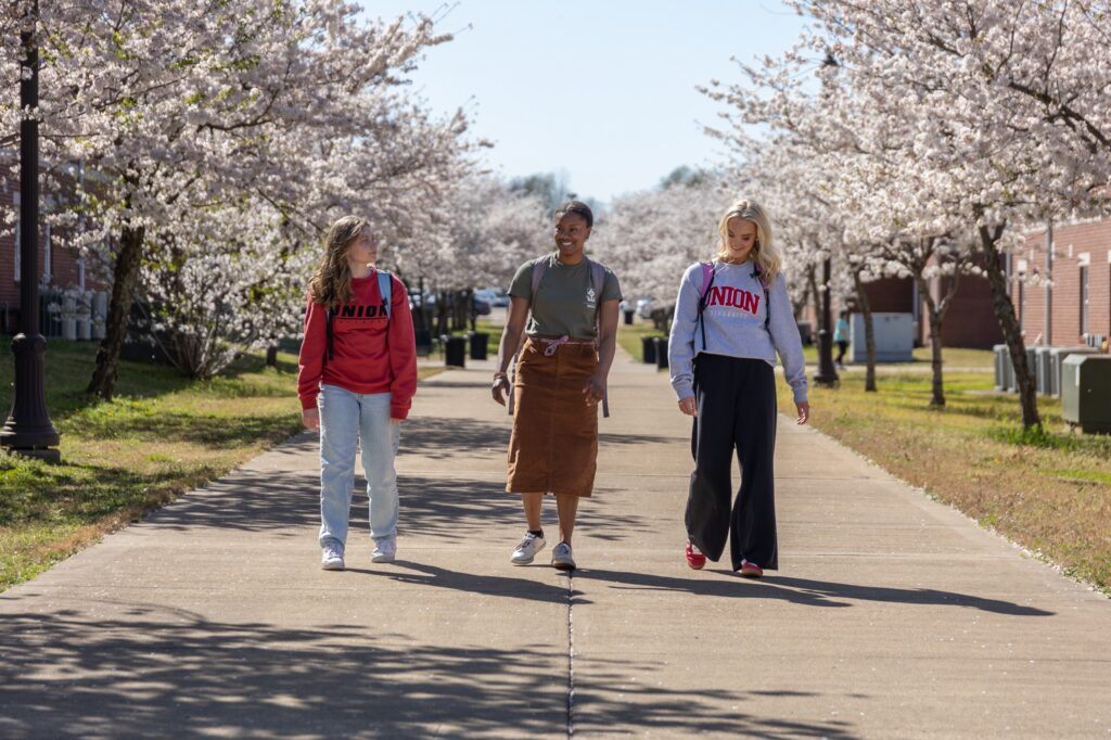 Three people walk down a wide sidewalk lined with blooming trees on both sides. Two wear UNION sweatshirts, and one wears a brown skirt with a green shirt, showcasing a vibrant 00-Component of campus life. Buildings and grass are visible in the background.