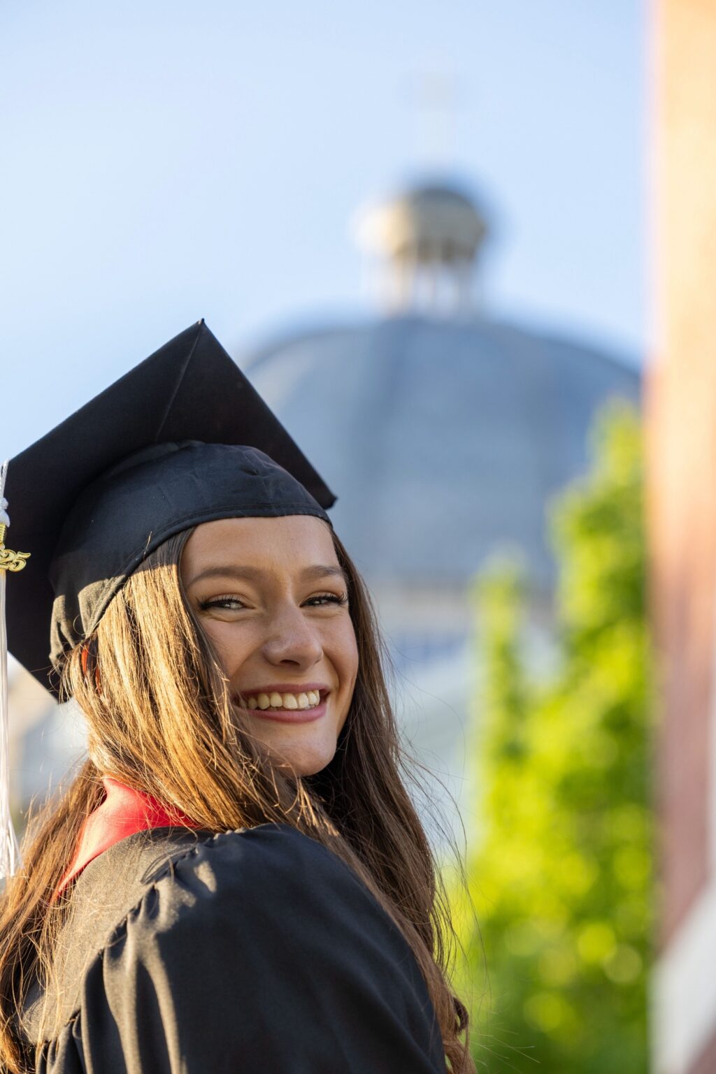 A young woman wearing a black graduation cap and gown smiles at the camera outdoors, with a domed building and green trees blurred in the background, celebrating her success as a proud member of the graduating class.