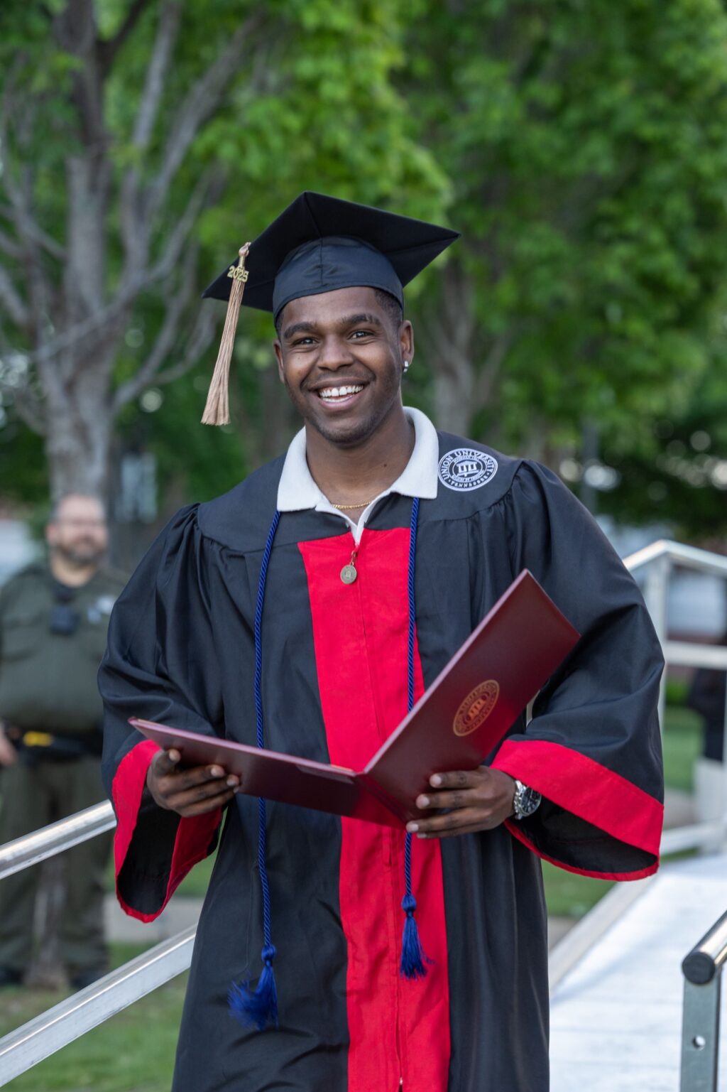 A graduate in a black and red cap and gown, proudly holding a diploma holder, smiles outdoors. The cap's red tassel adds a striking component to the scene, with green trees and a person in the background.