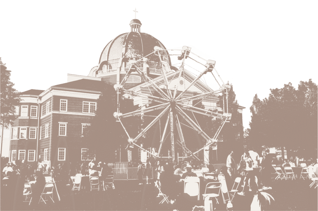 A Ferris wheel is set up on a lawn in front of a large domed building. People are seated at tables and walking around the area during what appears to be an outdoor event or festival.