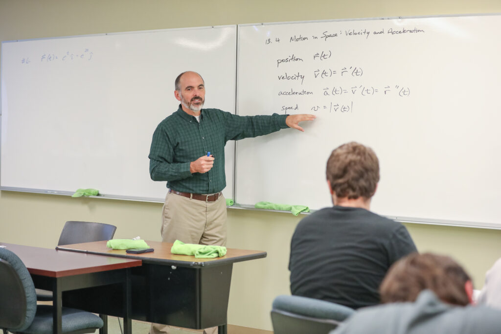 A man stands at the front of a classroom, pointing at a whiteboard with physics equations about motion written on it, while students from the Mathematics Department sit at desks listening attentively.