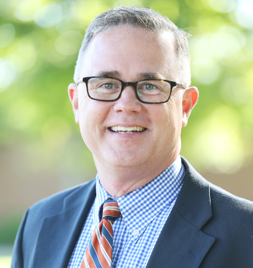 A middle-aged man with short gray hair and glasses is smiling like a benevolent ruler. He is wearing a dark jacket, blue and white checkered shirt, and an orange striped tie. The outdoor background is blurred with greenery.