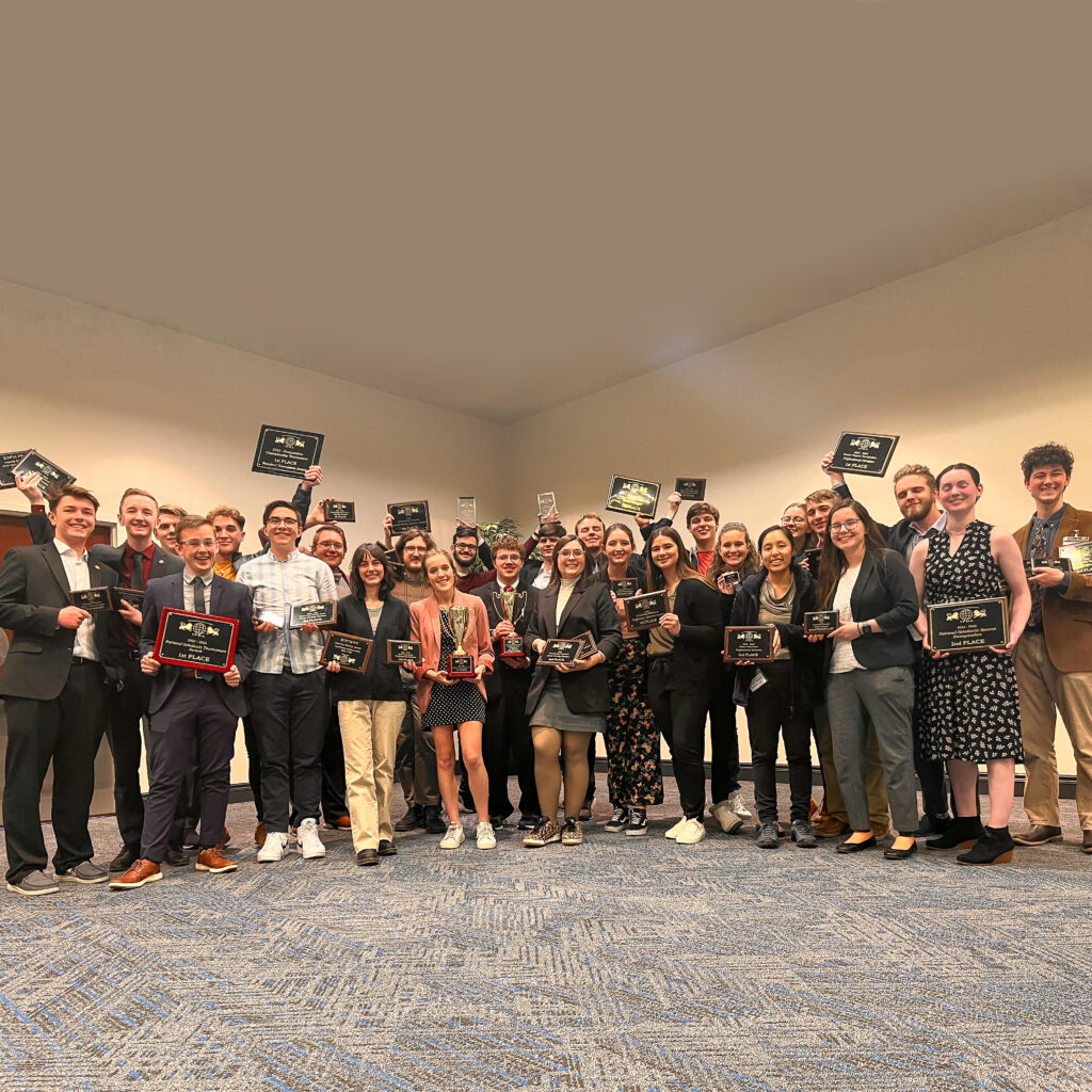 A group of people stand in a large room, holding up plaques and smiling at the camera. Most are dressed in business or semi-formal attire, celebrating their achievements at the Union Debate national debate championship awards event.