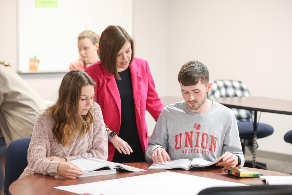 A woman in a pink blazer stands between a seated young woman and man, helping them with open books on a table—a scene reflecting dedicated teacher training at the School of Education. The man wears a Union University shirt; another person is visible in the background.