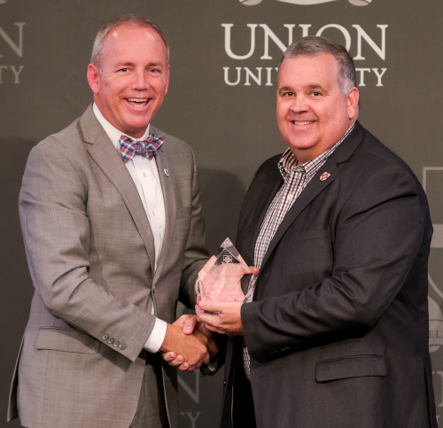 Two men in suits are smiling and shaking hands in front of a Union University backdrop. One man, named Union’s Alumnus of the Year during Homecoming week, is proudly holding a glass award.