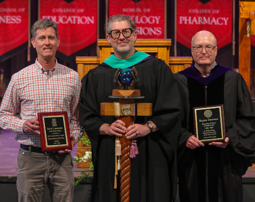 Three men stand indoors; the man in the center wears academic regalia and holds a ceremonial staff. Union students or faculty on either side hold plaques, with red Awards Day banners featuring school names hanging in the background.