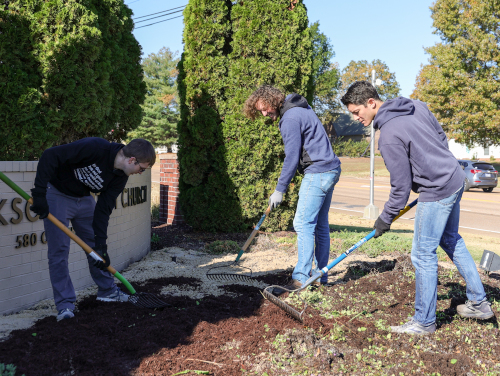 Three people wearing hoodies and gloves are using rakes to spread mulch and soil in a garden bed next to a brick sign and tall evergreen shrubs during Union's Campus and Community Day on a clear day.