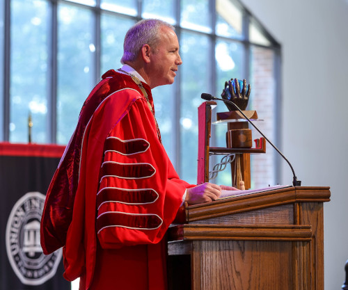 A man in a red academic robe speaks at a wooden podium indoors, addressing cultural shifts. Behind him is a black and red banner and a large window. On the podium sits a decorative object, possibly a trophy or sculpture.