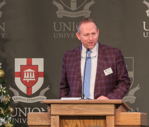 A man in a plaid jacket and tie speaks at a wooden podium in front of a Union University backdrop, addressing faculty and union trustees. A Christmas tree and the university emblem are visible in the background.
