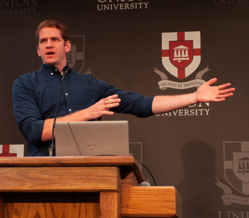A man stands behind a wooden podium with a laptop, gesturing with one arm extended. The backdrop displays Union University's logo and motto, Religio et Eruditio, at a youth apologetics conference.