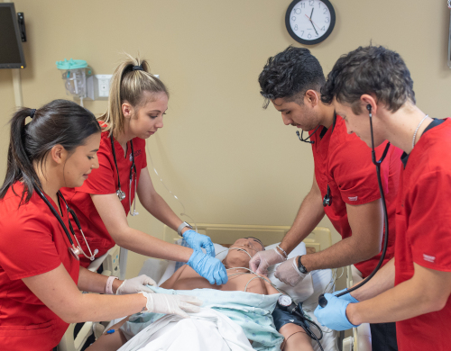 Four medical professionals in red scrubs, possibly from Vol State or Union University Hendersonville, attend to a patient in a hospital bed. Two use stethoscopes, one checks monitors, and another adjusts equipment. A clock is visible on the wall behind them.
