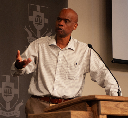 A man in a striped shirt stands at a wooden podium, speaking and gesturing with his right hand during the Jones Lecture. A microphone is attached to the podium, and a gray backdrop with a shield emblem is visible behind him.