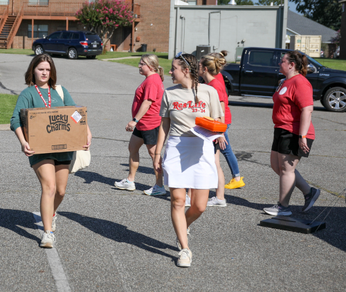 A group of five women, possibly new students, walks across a parking lot on Move-In Day. One woman carries a large box labeled Lucky Charms, while others have casual clothes and items. The Union, cars, and greenery are visible in the background.