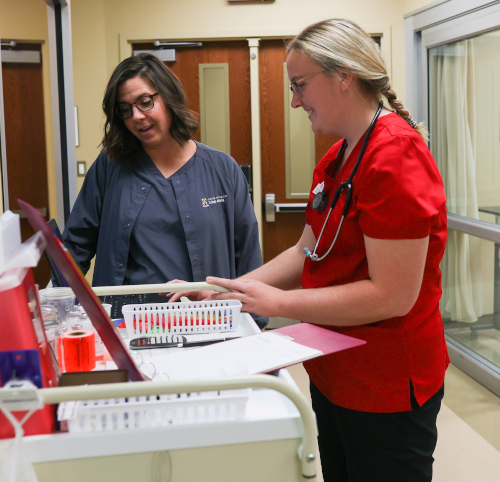 Two healthcare professionals stand at a medical cart, reviewing documents and supplies. Focused and engaged, they discuss strategies that help nursing grads achieve a perfect pass rate on the national licensure exam.
