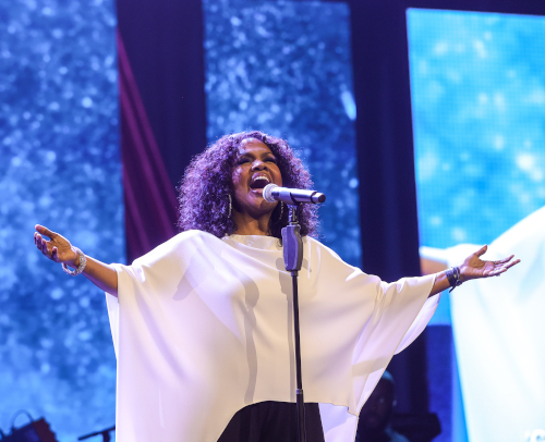 A woman with curly hair wearing a white flowing top sings passionately into a microphone on stage at the Union Scholarship Banquet, her arms outstretched as blue-toned screens and stage lights glow in the background.