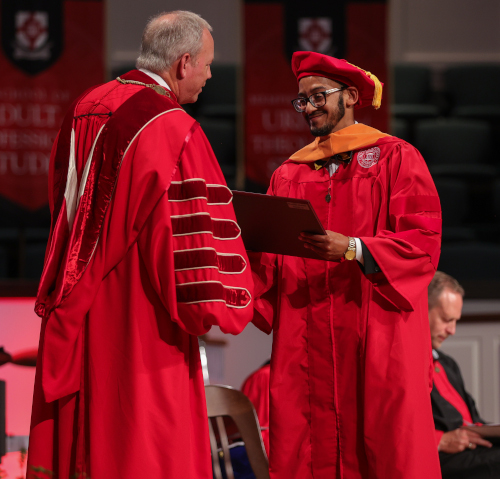 During a summer commencement, a student in red graduation attire receives a diploma from a faculty member in similar academic robes. Another person sits in the background as red banners hang on the wall, highlighting the graduation ceremony.