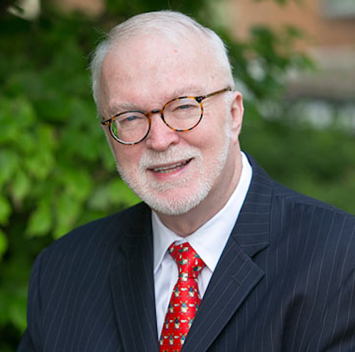 An older man with short white hair, a trimmed white beard, and glasses is wearing a dark pinstripe suit, white shirt, and red tie. He is smiling and standing outdoors in front of green foliage at the George Dockery Lectures on March 20.