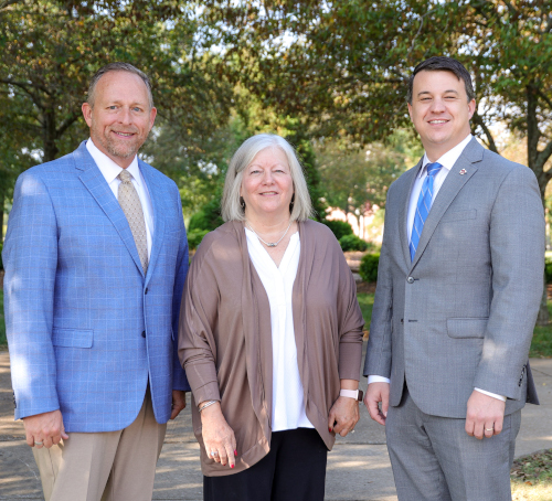 Three adults stand outdoors in business attire, posing for a photo. Among them are Union trustees, including the Board chair Perkins, dressed in suits and a taupe cardigan. Trees and greenery provide a lush background.