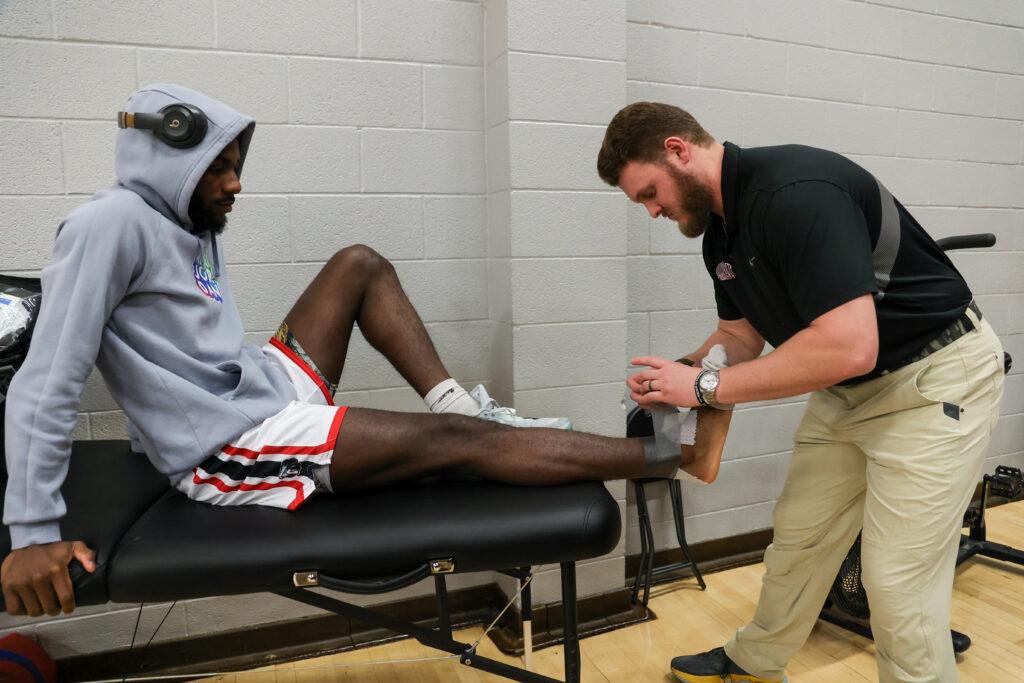 A man in athletic wear sits on a treatment table while another man, possibly a trainer from the Department of Exercise Science, wraps his left ankle with athletic tape in a gym setting.