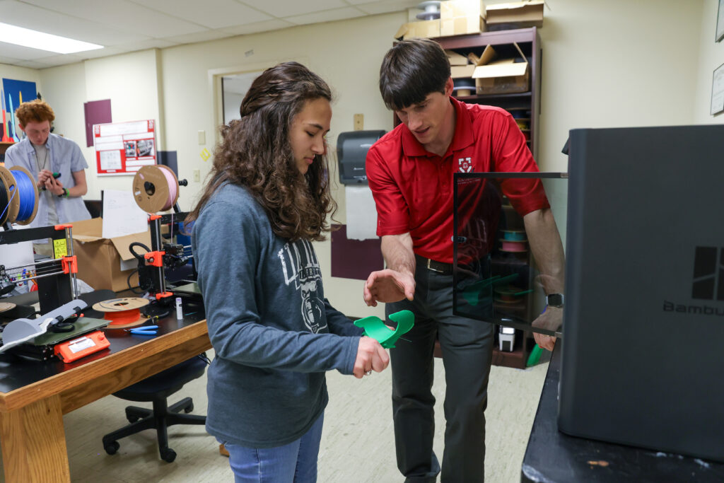 A man in a red shirt explains something to a young woman holding a green 3D-printed object in an Engineering department workshop. Another person works at a desk in the background with spools of filament and equipment.