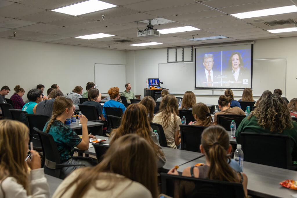 A classroom of students in a Political Science course sits facing a large screen showing a televised debate between two politicians. The students, seated at desks and some eating and drinking, watch the event attentively.