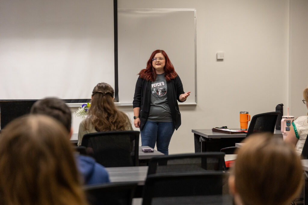 A woman with red hair stands at the front of a Psychology Department classroom, speaking to seated students. She wears jeans, a t-shirt, and a jacket. A phone and tumbler rest on a desk nearby. A whiteboard is behind her.