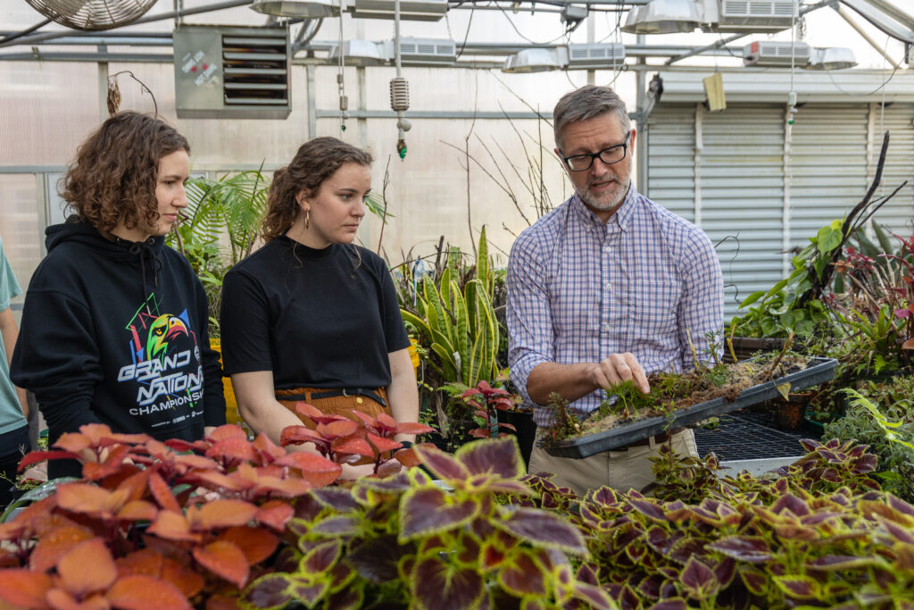A man in glasses and a checked shirt shows a tray of seedlings to two young women in a greenhouse filled with various plants, as part of a Biology Department project. The women are attentively observing the tray.