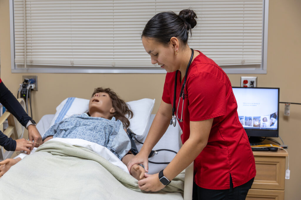 A nurse in a red uniform from the College of Nursing practices patient care with a medical mannequin lying in a hospital bed, while another person's hands adjust the blanket. A computer screen is visible on a nearby table.