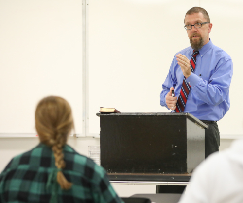 A man in a blue shirt and striped tie stands behind a podium, speaking to two seated individuals in a classroom with a whiteboard in the background at the Sept. 7 Youth Apologetics Conference EQUIP session.