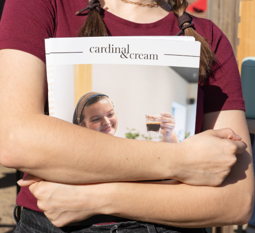 A person in a burgundy shirt with pigtail braids holds the Cardinal & Cream magazine, which highlights a BCA student competition and features a smiling woman raising a glass on its cover.