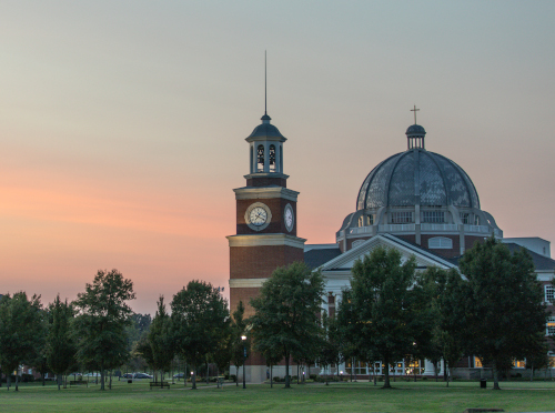 At sunset, Union’s large building with a clock tower and domed roof stands against soft pink and orange hues. The lawn in front is dotted with trees and benches, reflecting the classic charm of NCAA D2 schools.