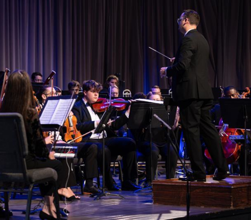 A conductor leads an orchestra of musicians from the music department, playing string and brass instruments on a stage, with sheet music visible and curtains in the background, setting the scene for memorable concerts.