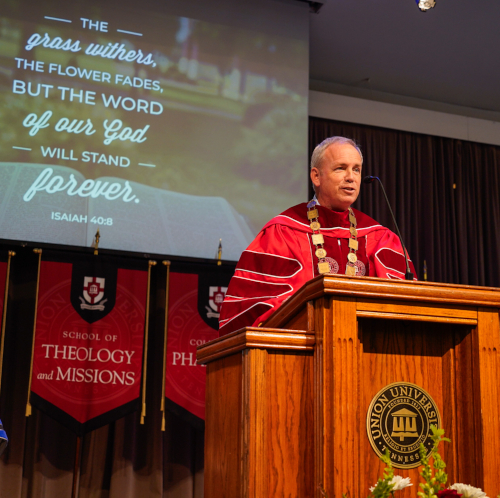 A man in academic regalia speaks with authority at a Union University podium. Behind him, a screen displays Scripture from Isaiah 40:8 and banners for the School of Theology and Missions and School of Pharmacy.