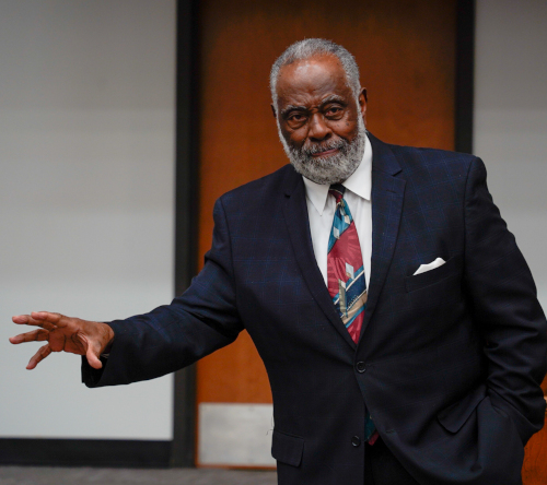 An older man with a gray beard, wearing a dark suit, white shirt, and patterned tie stands indoors near a wooden door, gesturing with his left hand as he speaks during the annual Dockery Lectures.