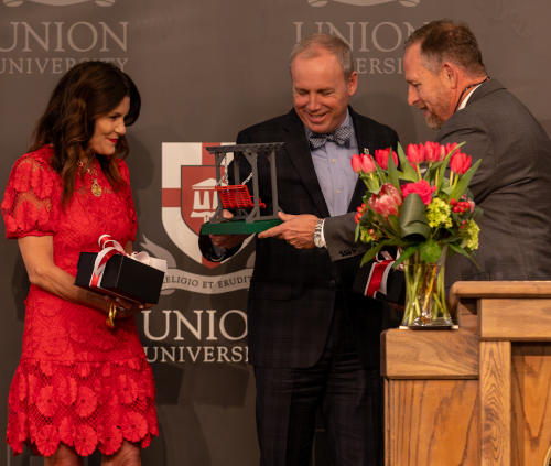 Three people stand before a Union University backdrop. During Oliver’s 10-year anniversary, one man hands a decorative object to another, while a woman in red holds a gift box. A bouquet of tulips sits on the podium nearby.