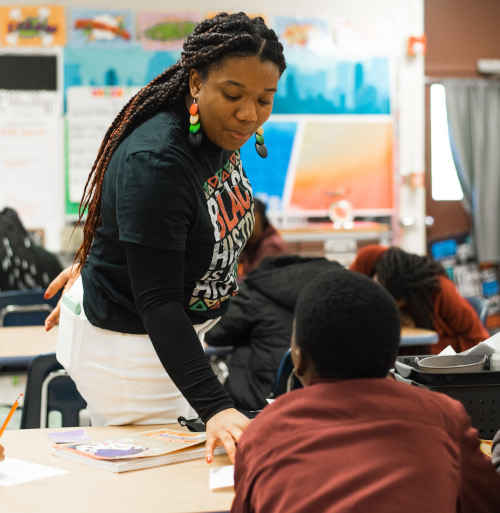 A teacher wearing a Black History shirt from the Union School of Education leans over to assist a student seated at a desk in a classroom, where other students work amid colorful posters and a screen displaying education scores in the background.
