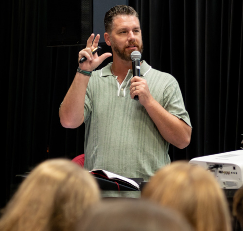 A man with a beard in a light green polo shirt speaks into a microphone while gesturing from behind a podium at the EQUIP apologetics conference, addressing an audience in a room with black curtains.