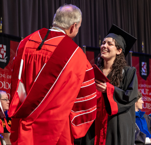A Union graduate in a cap and gown smiles while receiving her diploma from a faculty member in academic regalia during a graduation ceremony, celebrating faithful service and impact for Christ. Other people are seated in the background.